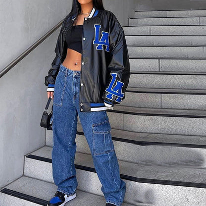 Frau in schwarzer LA-Baseballjacke, blauer Baggy-Jeans und Sneakers auf Treppe. Streetwear, urbaner Stil, modische Freizeitkleidung.