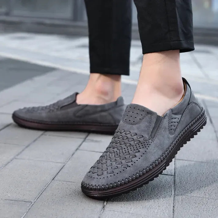 Graue Herren-Slipper aus Wildleder mit geflochtenem Design, rutschfeste Sohle, getragen auf Gehweg. Elegante Freizeitschuhe für Männer.