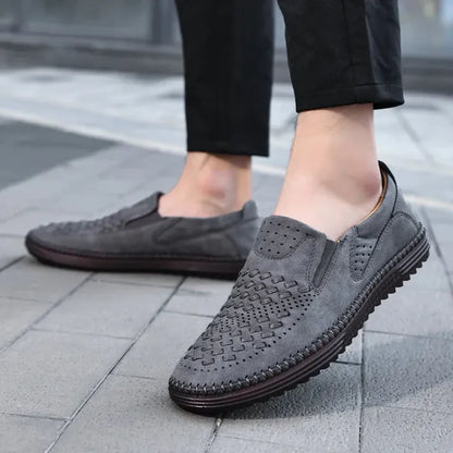 Graue Herren-Slipper aus Wildleder mit geflochtenem Design, rutschfeste Sohle, getragen auf Gehweg. Elegante Freizeitschuhe für Männer.