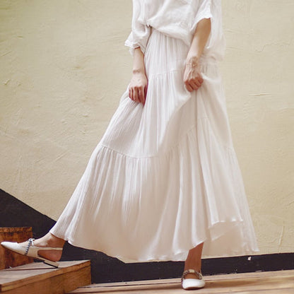 Frau in weißem, fließendem Maxirock und weißer Bluse, steht auf Holztreppe. Elegante Mode, Sommeroutfit, minimalistischer Stil.