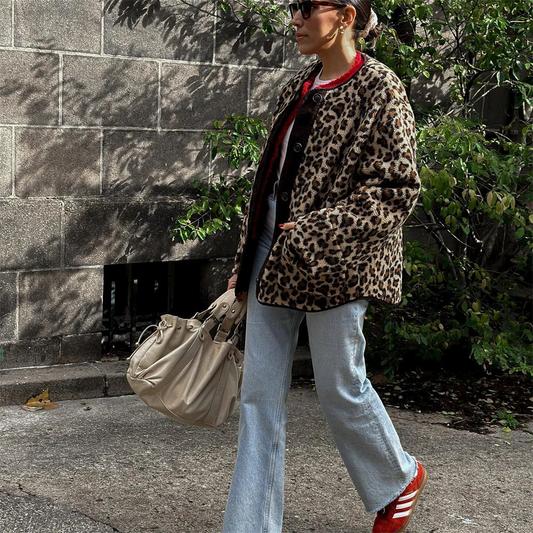 Frau in Leopardenmantel, blauer Jeans und roten Sneakers, trägt beige Handtasche, spaziert vor grauer Wand und grünen Büschen. Streetstyle, Mode.