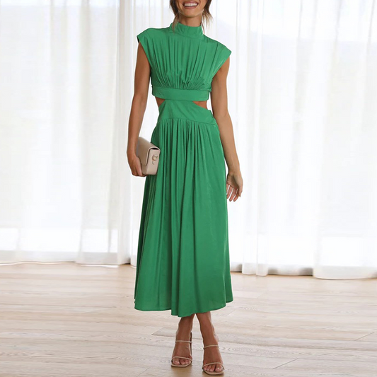 Grünes Maxikleid mit Cut-Outs, ärmellos, elegantes Design, Frau hält eine beige Clutch, steht auf Holzboden vor weißen Vorhängen.