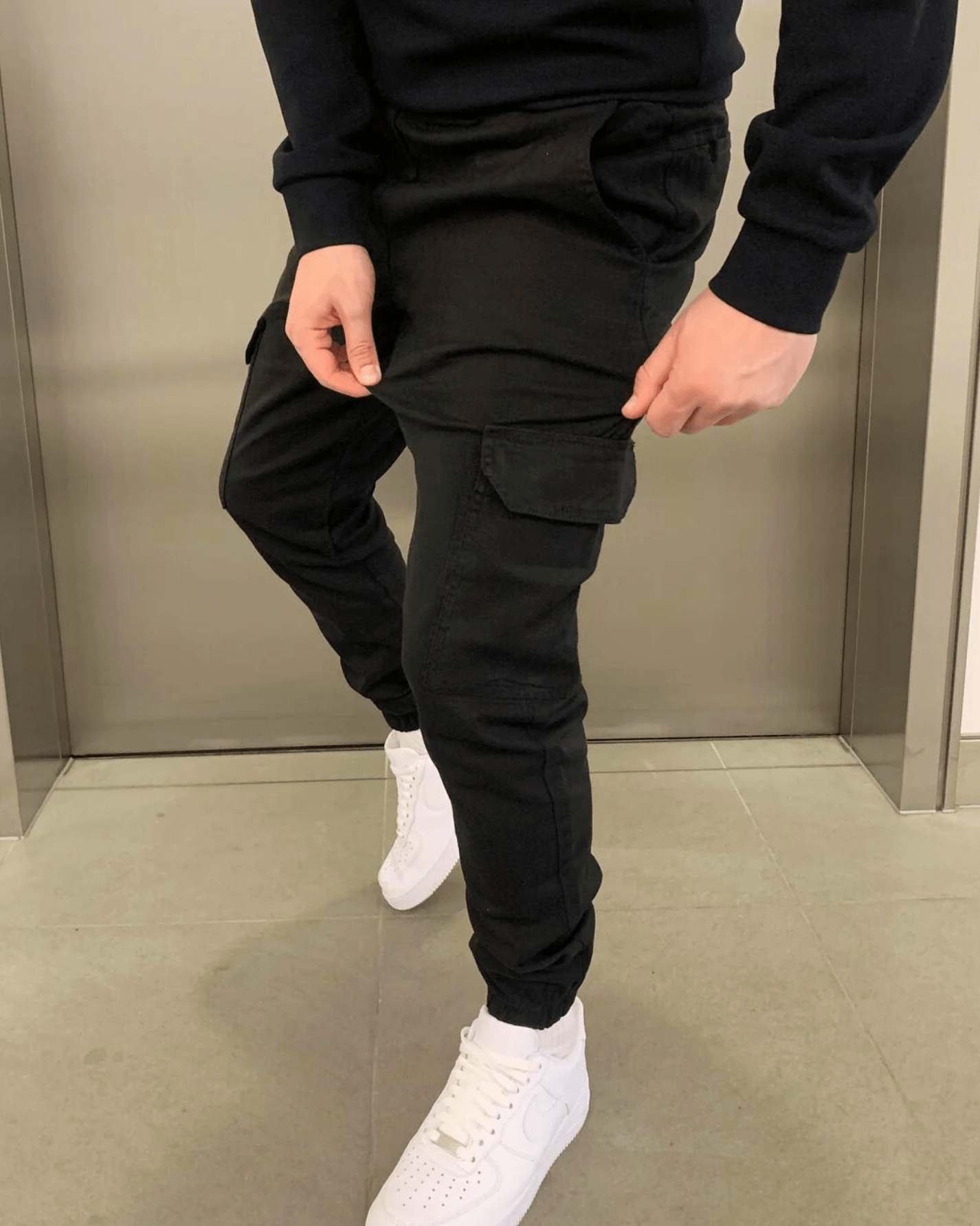 Schwarze Cargohose und weiße Sneaker, getragen von einer Person in einem Aufzug. Modetrend, lässiger Stil, Herrenbekleidung, urbane Mode.