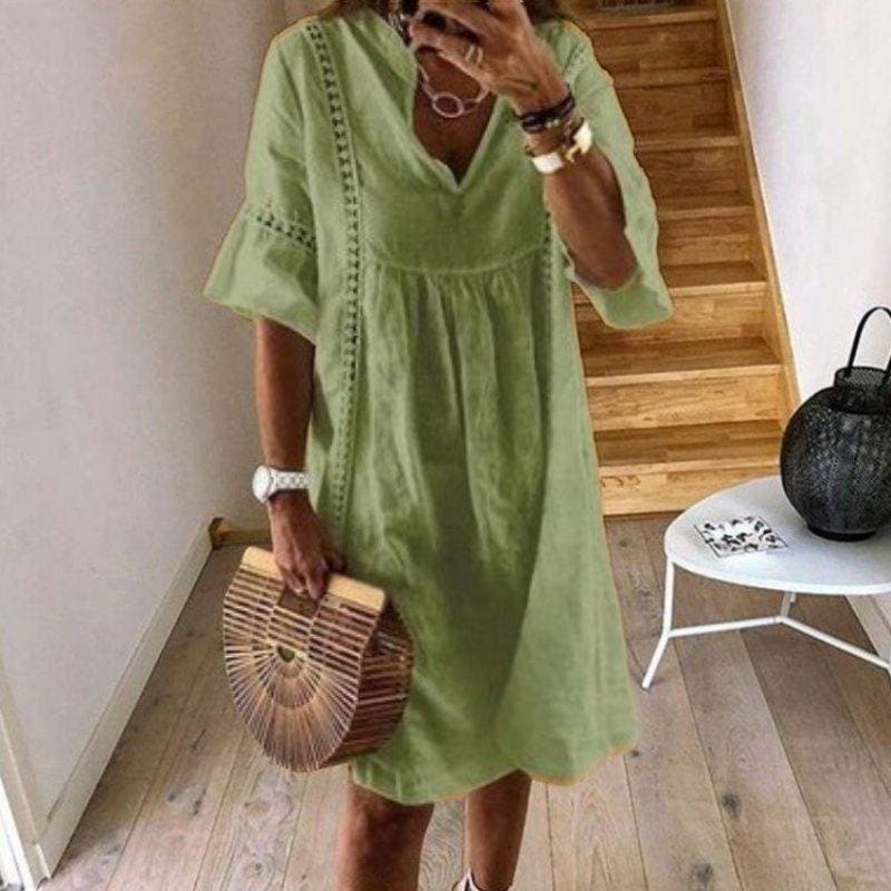 Frau in grünem Leinenkleid mit V-Ausschnitt, hält runde Bambustasche. Steht vor Holztreppe, moderne Einrichtung. Sommermode, Boho-Stil.