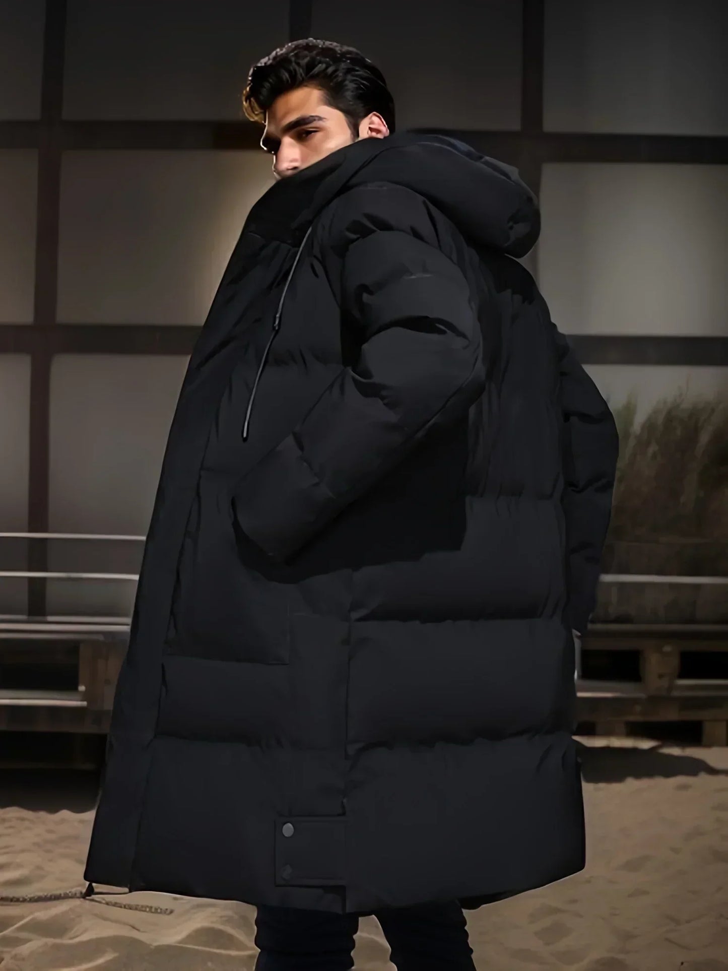Mann in schwarzem, übergroßem Daunenmantel mit Kapuze, steht seitlich auf Sandboden. Wintermode, Outdoor-Bekleidung, stilvoller Look.