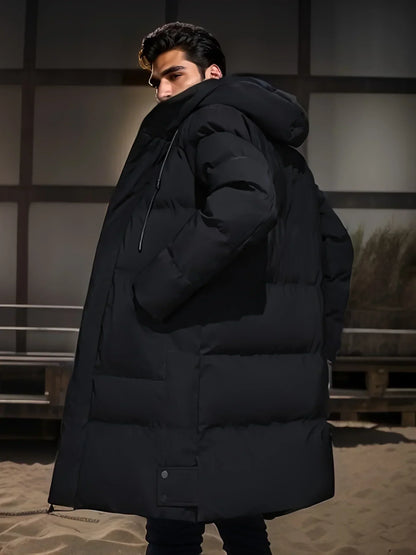 Mann in schwarzem, übergroßem Daunenmantel mit Kapuze, steht seitlich auf Sandboden. Wintermode, Outdoor-Bekleidung, stilvoller Look.