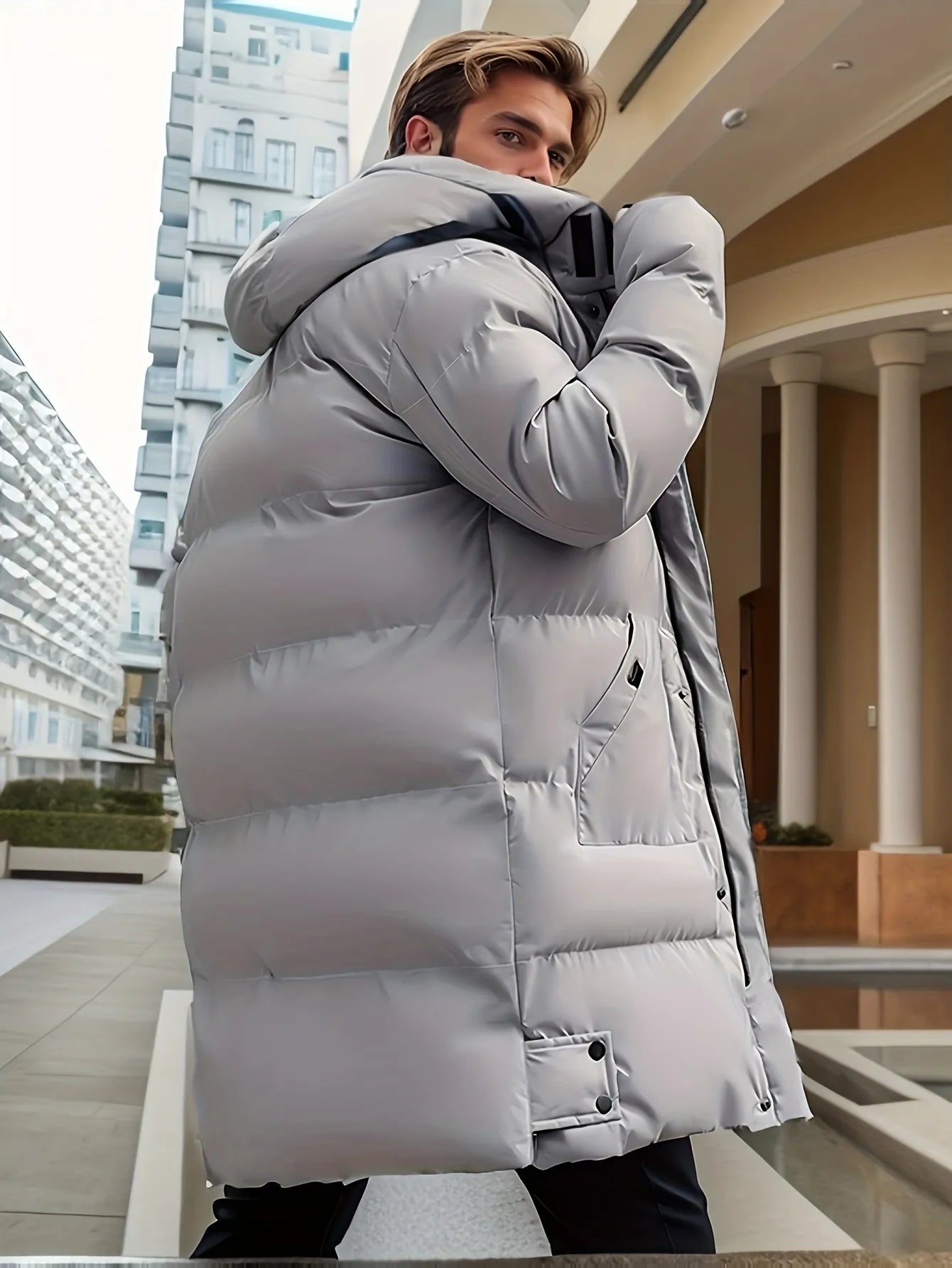 Mann in grauem Daunenmantel, moderne Architektur im Hintergrund, Wintermode, stilvolle Herrenbekleidung, urbane Umgebung.
