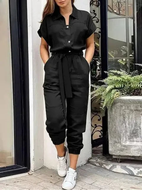 Frau in schwarzem Jumpsuit mit Gürtel, steht vor einem Fenster. Sie trägt weiße Sneaker. Moderne Damenmode, lässiger Stil, urbaner Look.
