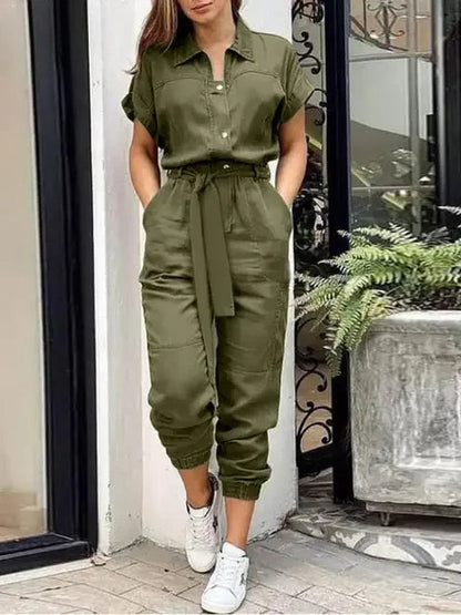 Frau in olivgrünem Jumpsuit mit Gürtel und weißen Sneakers steht vor einem modernen Gebäude. Modetrend, Damenmode, lässiger Stil.