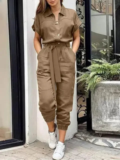 Frau in braunem Jumpsuit mit kurzen Ärmeln und Gürtel, kombiniert mit weißen Sneakers, steht vor einem modernen Gebäude mit Pflanzen.