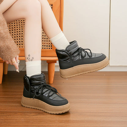 Schwarze Winterstiefel mit dicker Sohle, getragen von Person mit weißen Socken und tätowiertem Bein, auf Holzboden vor Korbstuhl.