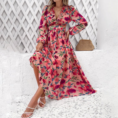Frau in langem, geblümtem Kleid vor weißem Gitter, hält Rocksaum. Sommermode, florales Design, eleganter Look, trendiges Maxikleid.