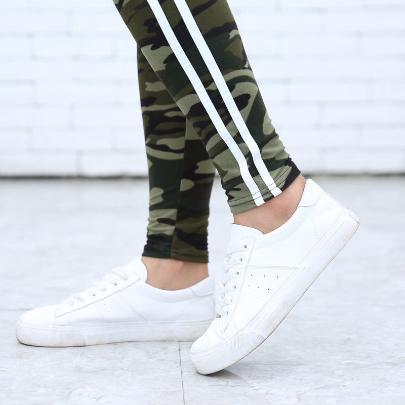 Weiße Sneaker und Camouflage-Leggings mit Streifen auf grauem Boden vor weißer Ziegelwand. Modetrend, Streetwear, Damenbekleidung.