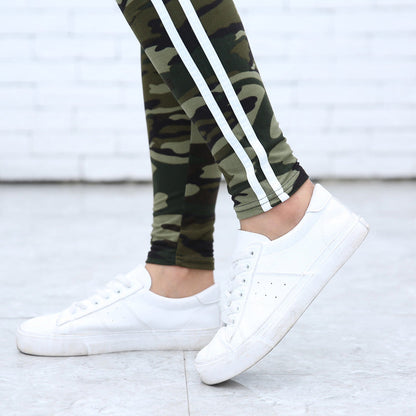 Weiße Sneaker und Camouflage-Leggings mit Streifen auf grauem Boden vor weißer Ziegelwand. Modetrend, Streetwear, Damenbekleidung.