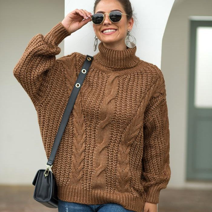 Frau in braunem Strickpullover mit Zopfmuster, Sonnenbrille und schwarzer Umhängetasche, lächelnd vor weißem Hintergrund. Modetrend Herbst 2023.