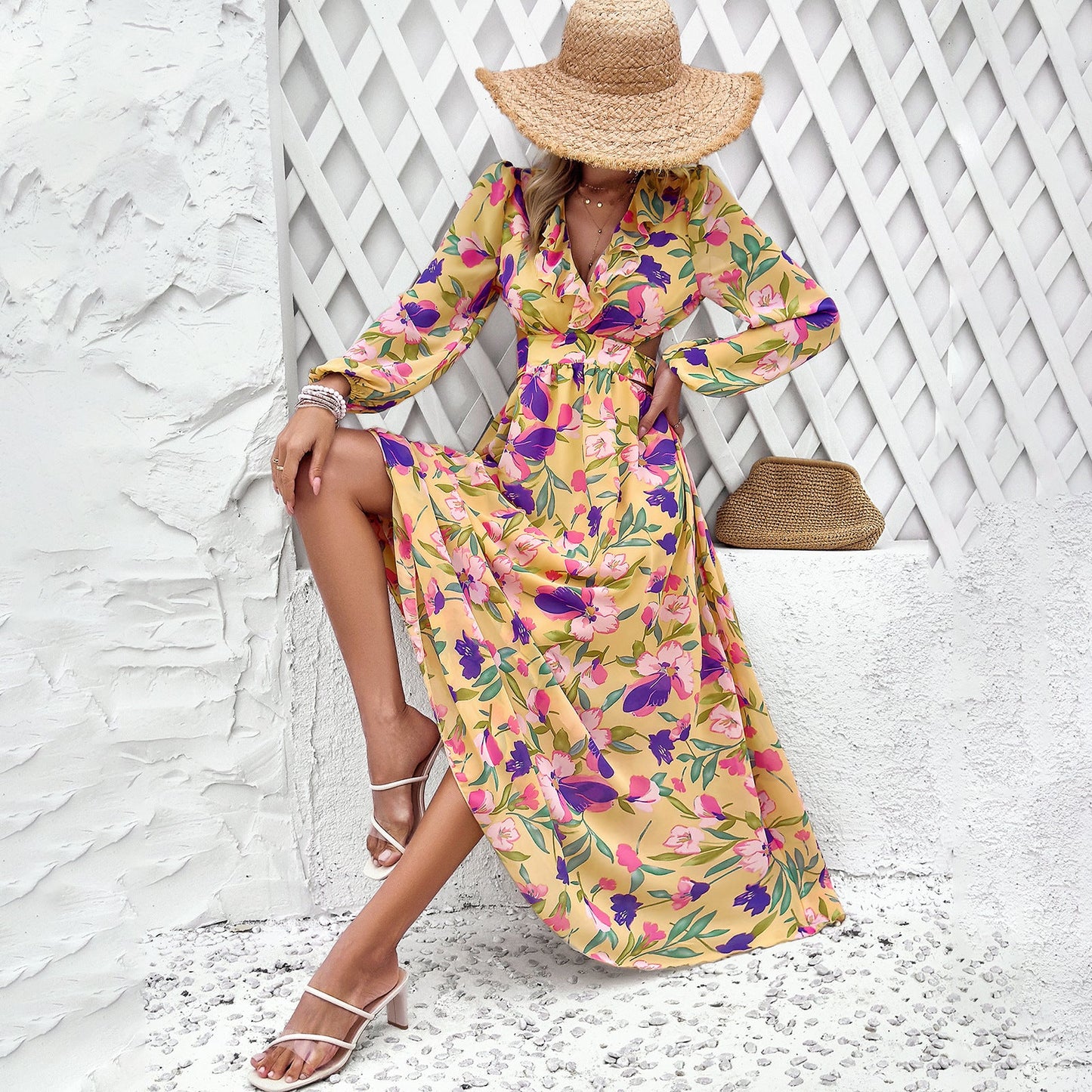 Frau in gelbem, geblümtem Maxikleid und Strohhut sitzt vor weißer Wand. Sommermode, elegantes Design, Freizeitkleidung, florales Muster.