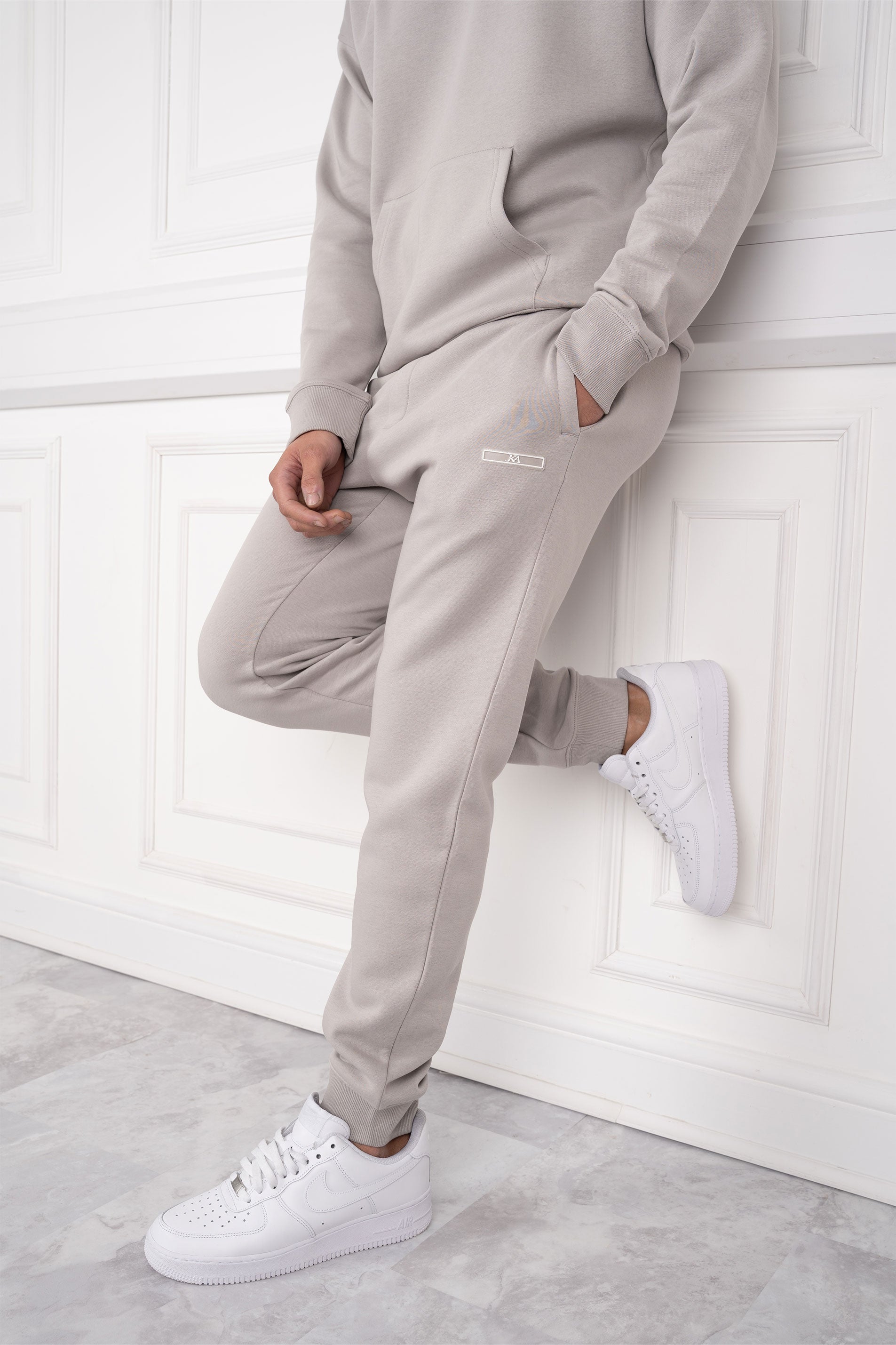 Mann in grauem Jogginganzug lehnt an weißer Wand, trägt weiße Sneaker. Bequeme Freizeitmode, sportlicher Look, Herrenbekleidung.