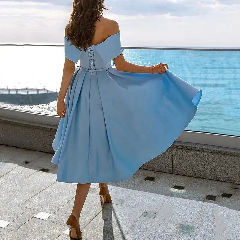 Frau in schulterfreiem, blauem Kleid mit Schnürung, blickt aufs Meer. Elegantes Sommeroutfit, perfekte Abendmode am Strand.