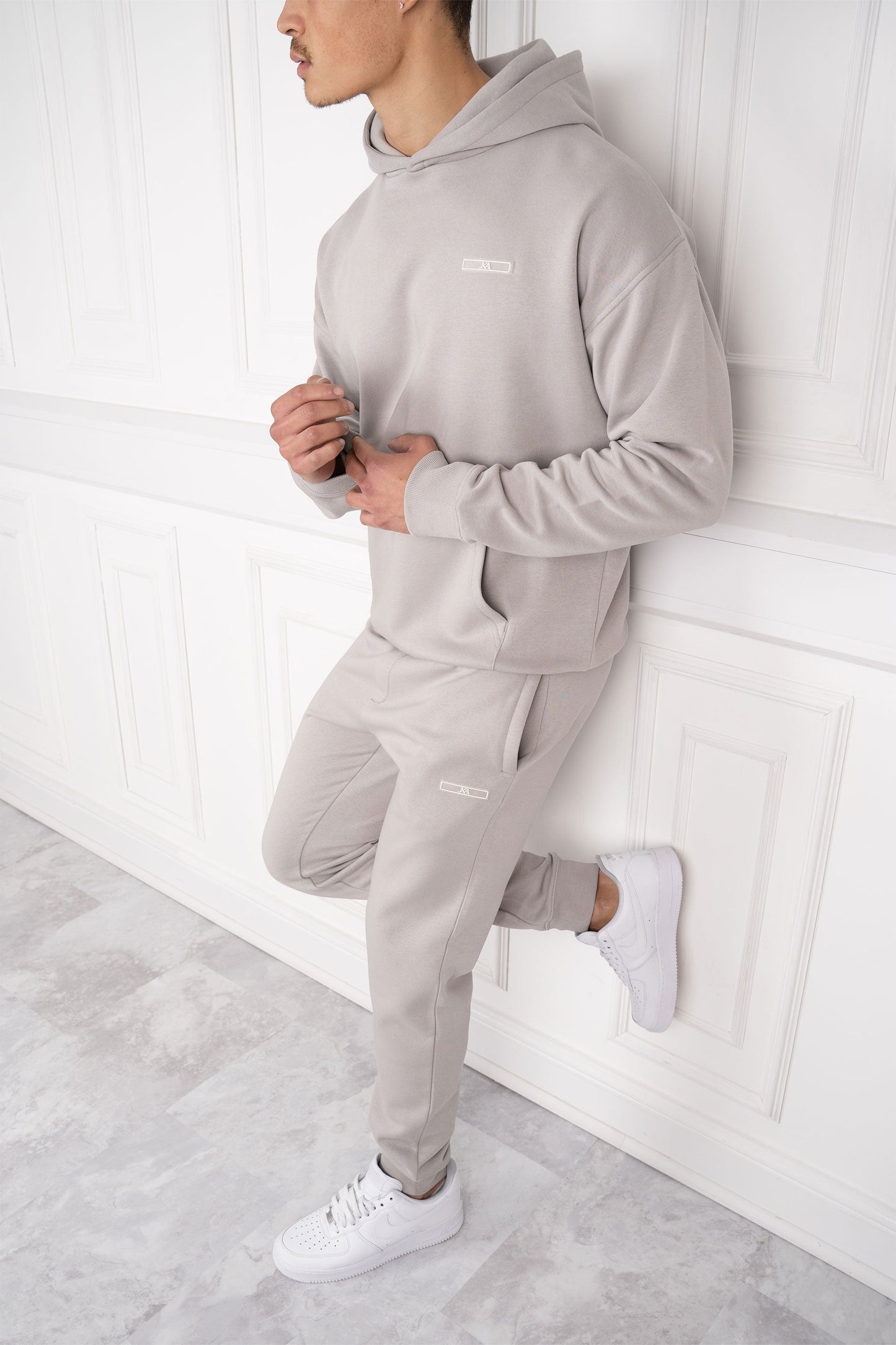 Mann in grauem Hoodie und Jogginghose lehnt an weißer Wand. Sportliche Freizeitmode, bequeme Kleidung, moderner Stil, Herrenmode.