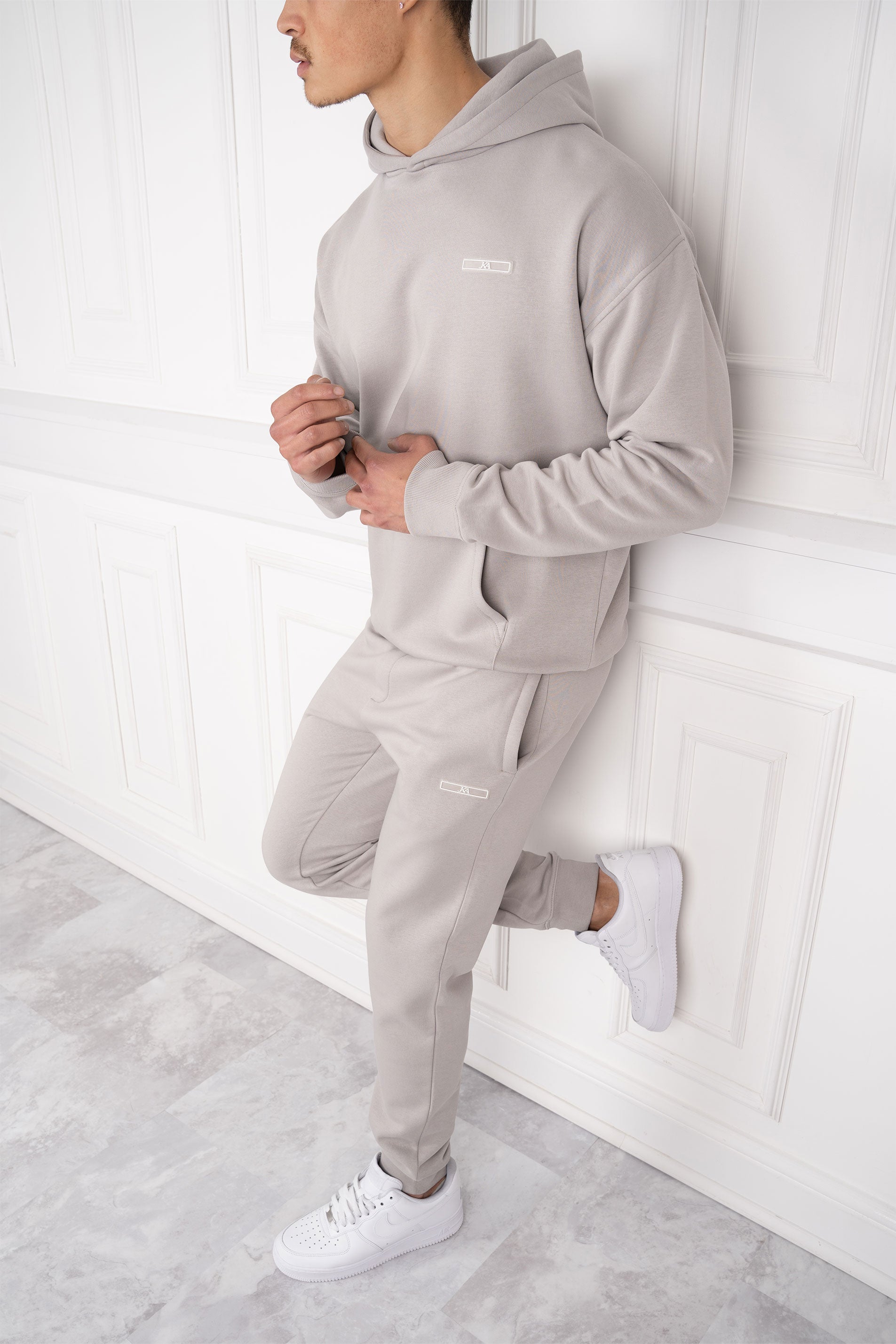 Mann in grauem Hoodie und Jogginghose lehnt an weißer Wand. Sportliche Freizeitmode, bequeme Kleidung, moderner Stil, Herrenmode.