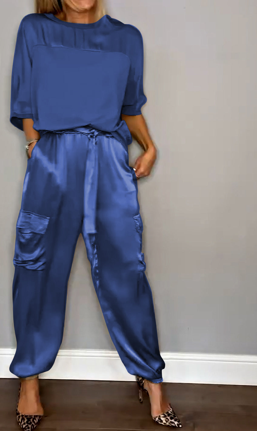 Frau in blauer Satin-Hose und passendem Oberteil, steht vor grauer Wand. Modetrend, elegante Damenbekleidung, stilvolle Freizeitmode.