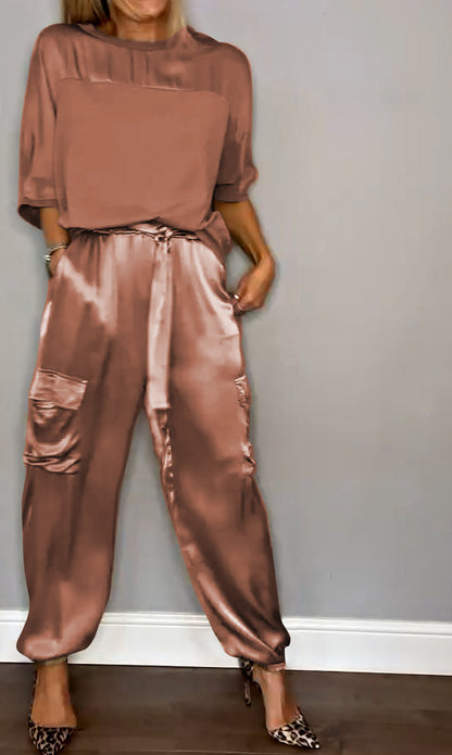 Frau in eleganter, rosafarbener Satin-Hose mit Taschen und passendem Oberteil, steht vor grauer Wand. Modetrend, Damenmode, stilvoll.