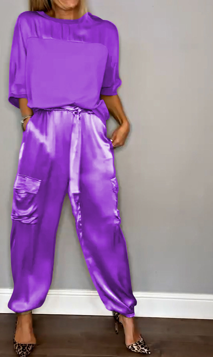 Frau in lila Satin-Overall mit Taschen, kombiniert mit Leoparden-High-Heels, vor grauer Wand. Modetrend, eleganter Look, Damenmode.