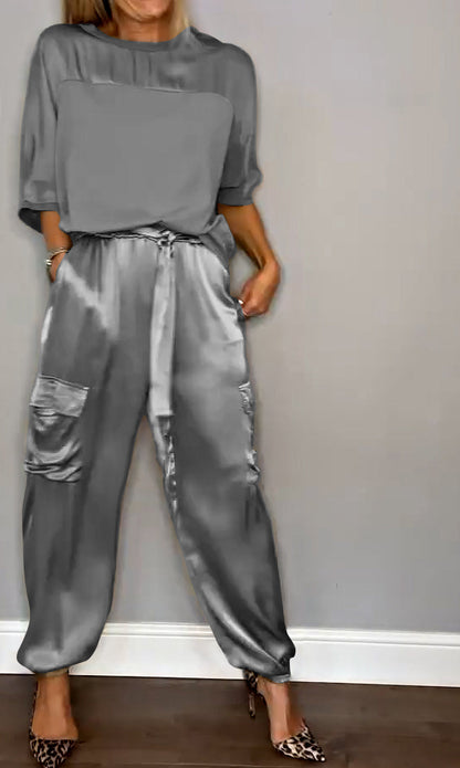 Frau in grauem Satin-Overall mit Taschen, steht vor grauer Wand, trägt Leoparden-High-Heels. Elegante Damenmode, stilvoller Look.