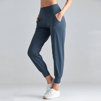 Frau in blauer Jogginghose und weißen Turnschuhen, seitlich stehend, Hände in den Taschen, sportliche Kleidung, Fitnessmode, grauer Hintergrund.
