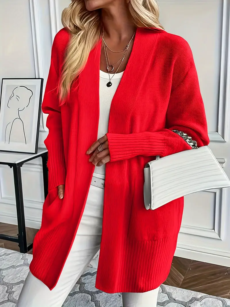 Frau in rotem Strickcardigan, weißem Top und Hose, hält weiße Handtasche. Mode, Herbstoutfit, eleganter Stil, Innenraum.