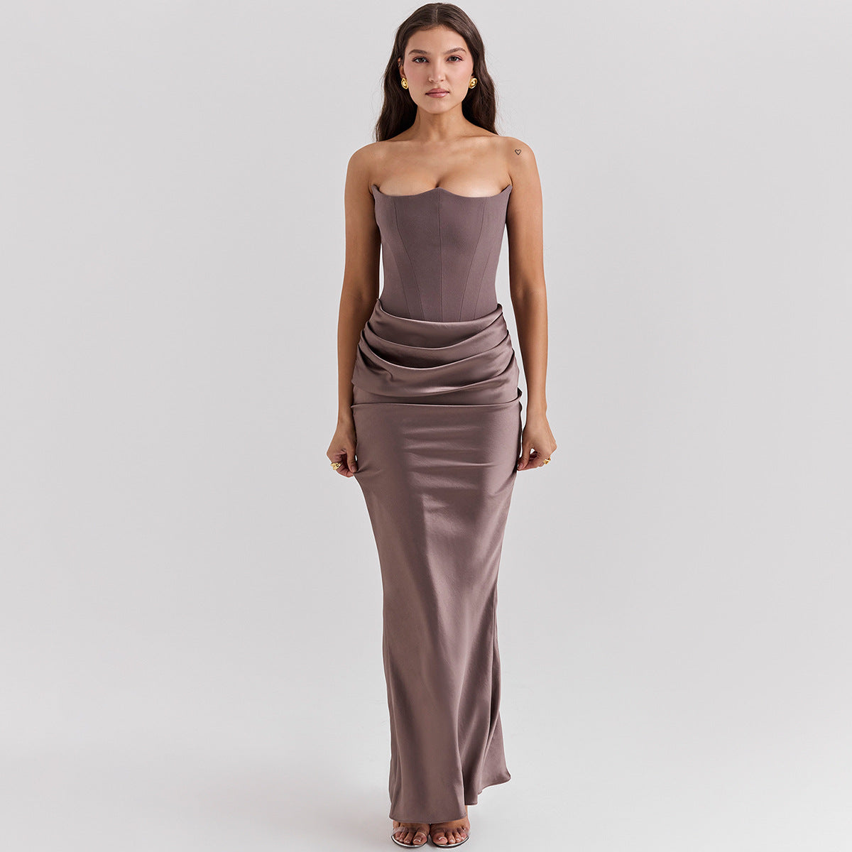 Frau in eleganter, trägerloser, taupefarbener Abendkleid mit drapiertem Rock, stehend vor neutralem Hintergrund. Perfekt für formelle Anlässe.