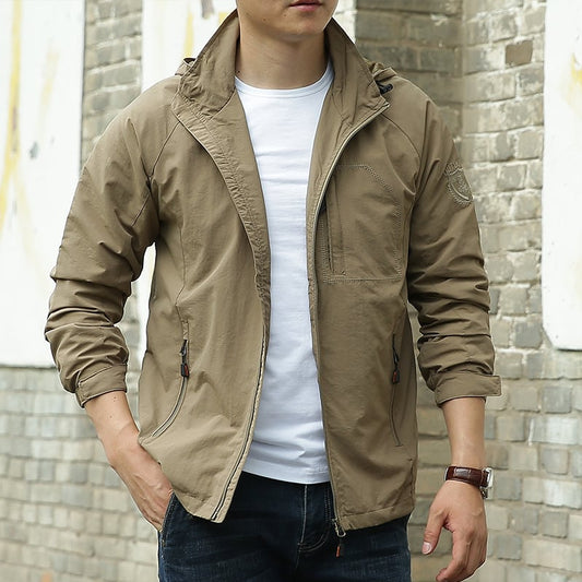 Mann in beiger Jacke und weißem T-Shirt vor Backsteinmauer, lässiger Stil, Mode für Herren, Outdoor-Bekleidung, Freizeitlook.