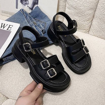 Schwarze Damen-Sandalen mit Blockabsatz und drei verstellbaren Schnallen auf beigem Sofa, neben blauer Jeans und Magazin.