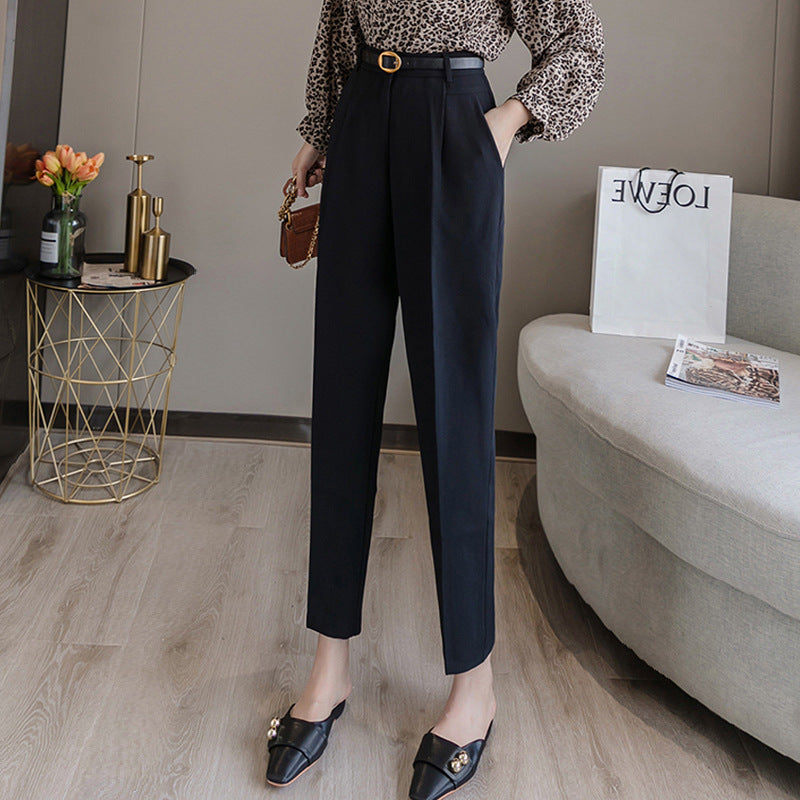 Frau in eleganter schwarzer Hose und gemusterter Bluse, steht in modernem Wohnzimmer mit Sofa, Blumen und Einkaufstasche. Mode, Damenbekleidung, Stil.