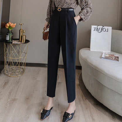 Frau in eleganter schwarzer Hose und gemusterter Bluse, steht in modernem Wohnzimmer mit Sofa, Blumen und Einkaufstasche. Mode, Damenbekleidung, Stil.