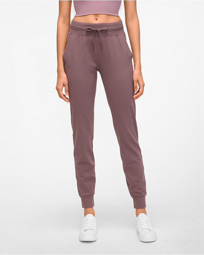 Frau in lila Jogginghose und weißem Top, stehend vor weißem Hintergrund. Bequeme Sportbekleidung, ideal für Fitness und Freizeit.