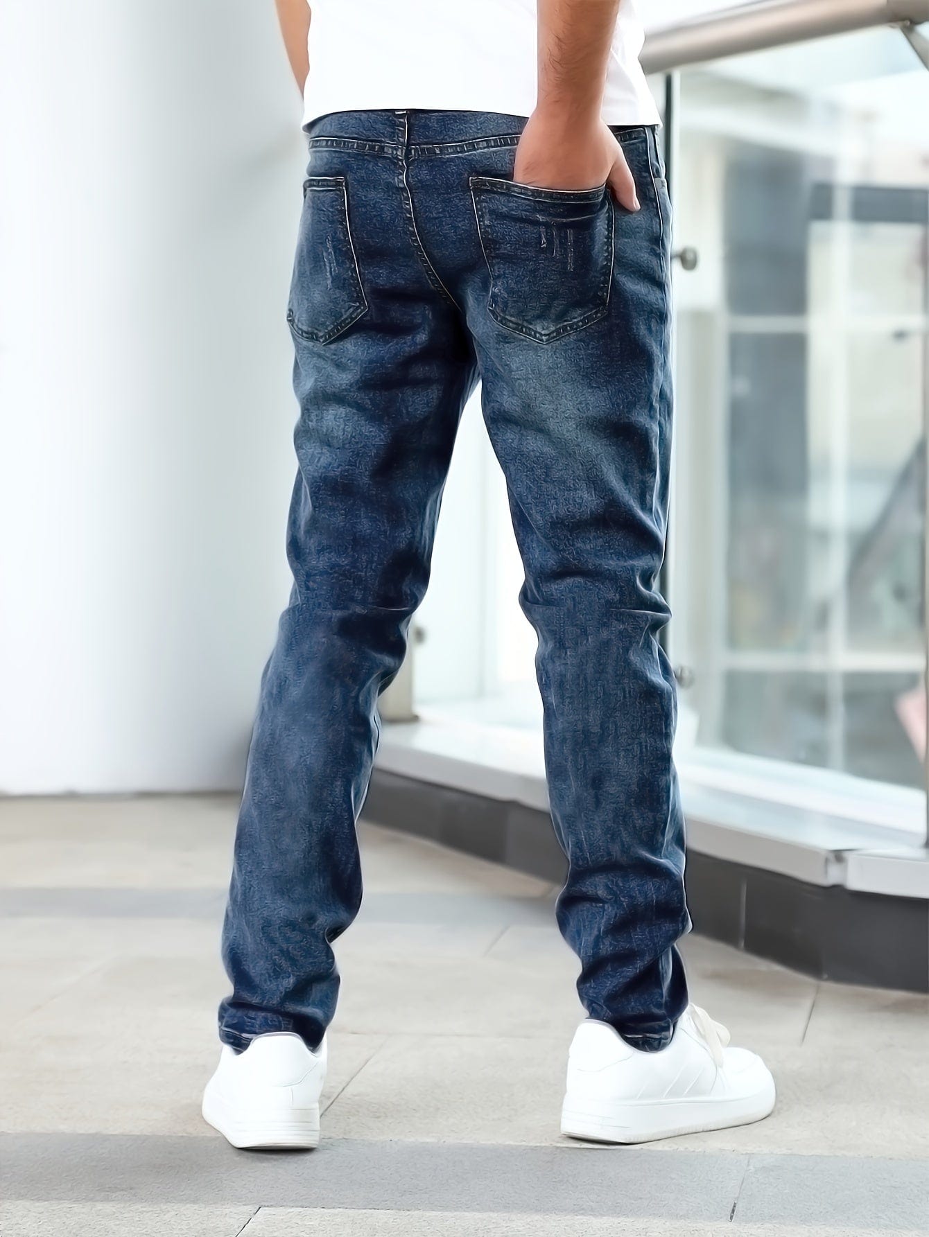 Mann in blauer Jeans und weißen Sneakern steht in modernem Gebäude. Herrenmode, Denim, lässiger Stil, Rückansicht.