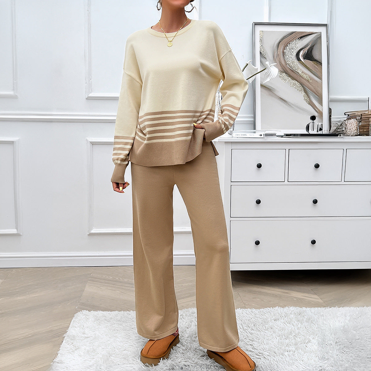 Frau in beige-weißem Strickpullover und Hose, steht in modernem Wohnzimmer mit weißem Teppich und Kommode. Elegante, gemütliche Damenmode.