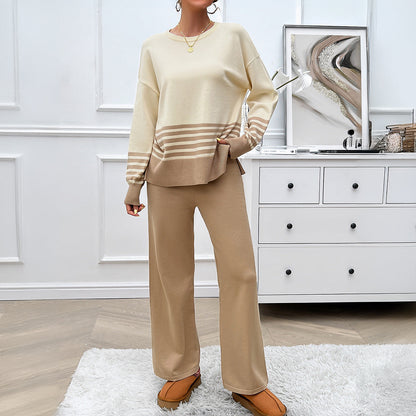 Frau in beige-weißem Strickpullover und Hose, steht in modernem Wohnzimmer mit weißem Teppich und Kommode. Elegante, gemütliche Damenmode.