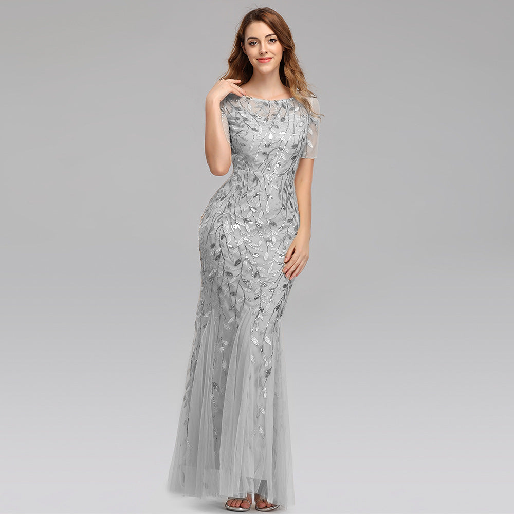 Frau in elegantem, bodenlangem, silbernem Abendkleid mit floralen Mustern, lächelnd, grauer Hintergrund. Perfekt für formelle Anlässe und Hochzeiten.