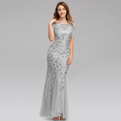 Frau in elegantem, bodenlangem, silbernem Abendkleid mit floralen Mustern, lächelnd, grauer Hintergrund. Perfekt für formelle Anlässe und Hochzeiten.