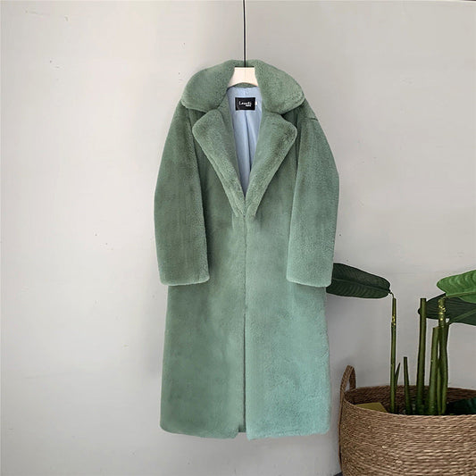 Grüner Damen-Wollmantel mit Reverskragen, hängend an weißer Wand, neben Korb mit Pflanzen. Eleganter Wintermantel, modisch und warm.