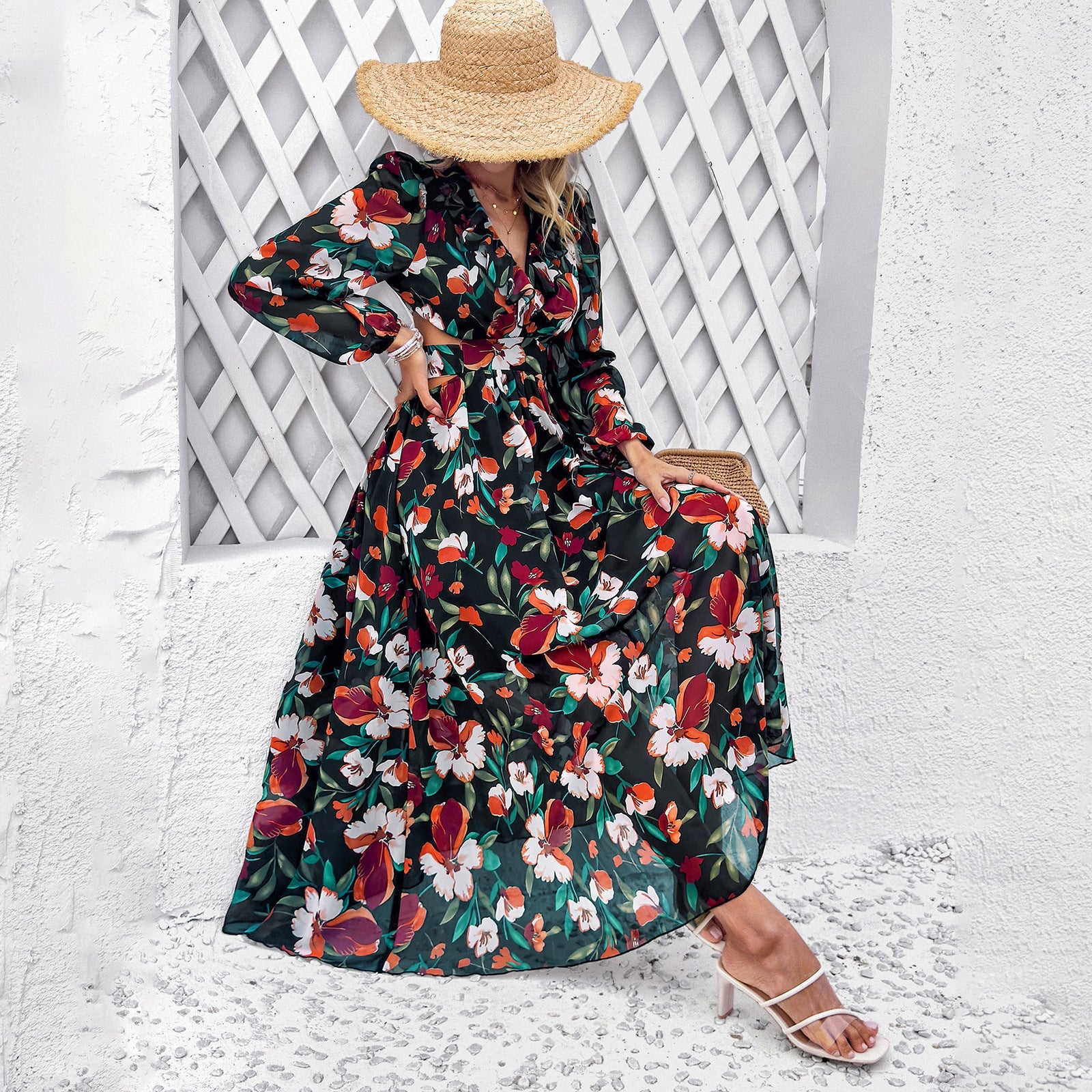 Frau in geblümtem Maxikleid und Strohhut sitzt vor weißer Wand. Sommermode, elegantes Kleid, florales Muster, stilvolle Sandalen.