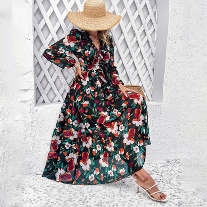 Frau in geblümtem Maxikleid und Strohhut sitzt vor weißer Wand. Sommermode, elegantes Kleid, florales Muster, stilvolle Sandalen.
