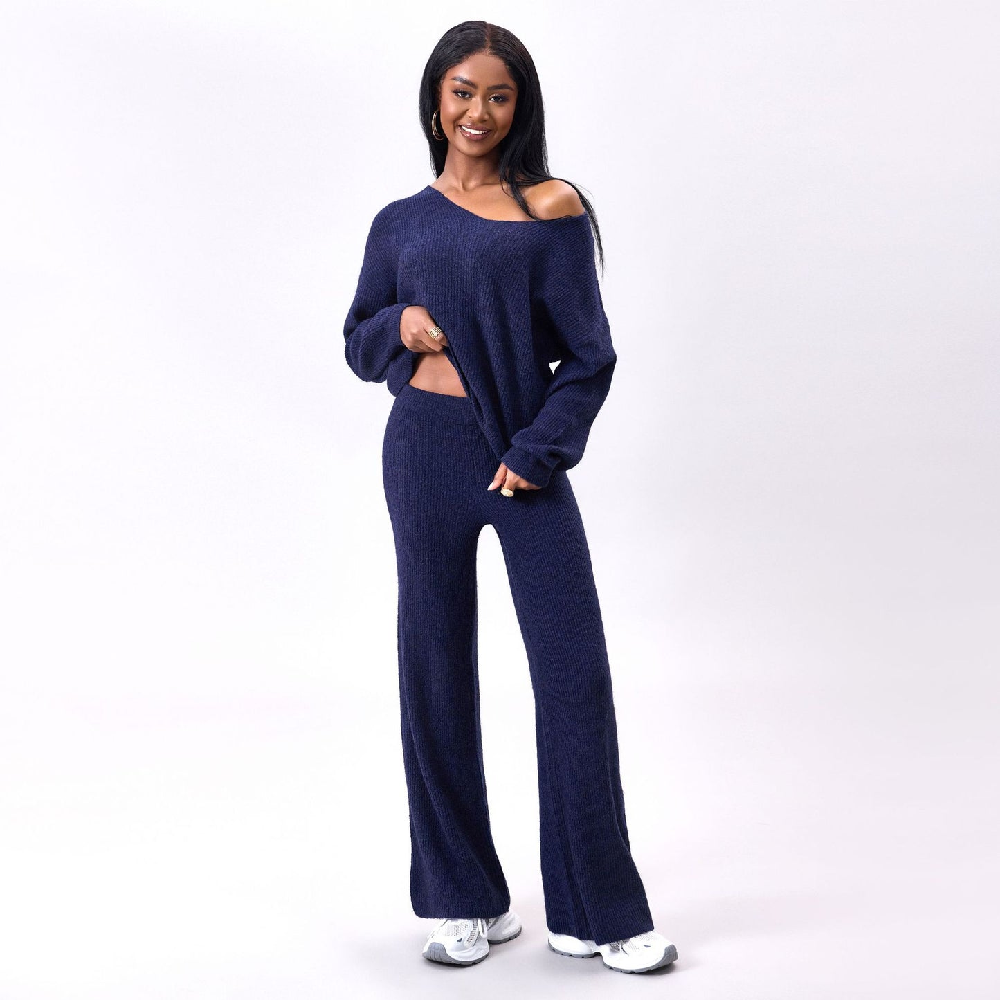 Frau in dunkelblauem Strick-Zweiteiler mit weitem Pullover und Hose, lächelt, trägt weiße Sneaker, modisches Freizeitoutfit, Studioaufnahme.