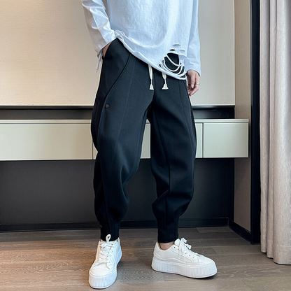 Mann trägt schwarze Jogginghose und weiße Sneakers, kombiniert mit zerrissenem weißen Langarmshirt. Moderne Herrenmode, lässiger Streetwear-Stil.