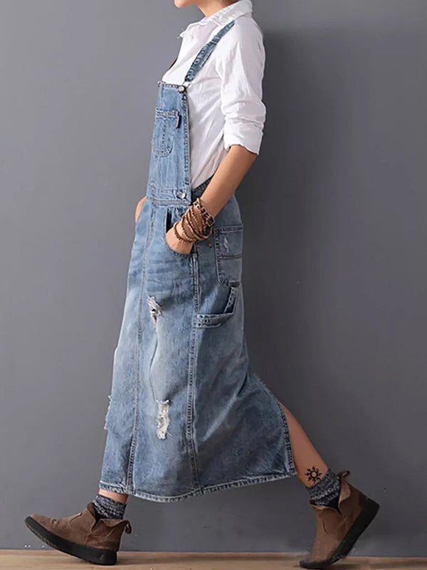 Frau in zerrissenem Jeans-Latzkleid, weißem Hemd und braunen Stiefeln vor grauer Wand. Modetrend, lässiger Stil, Denim-Kleid.