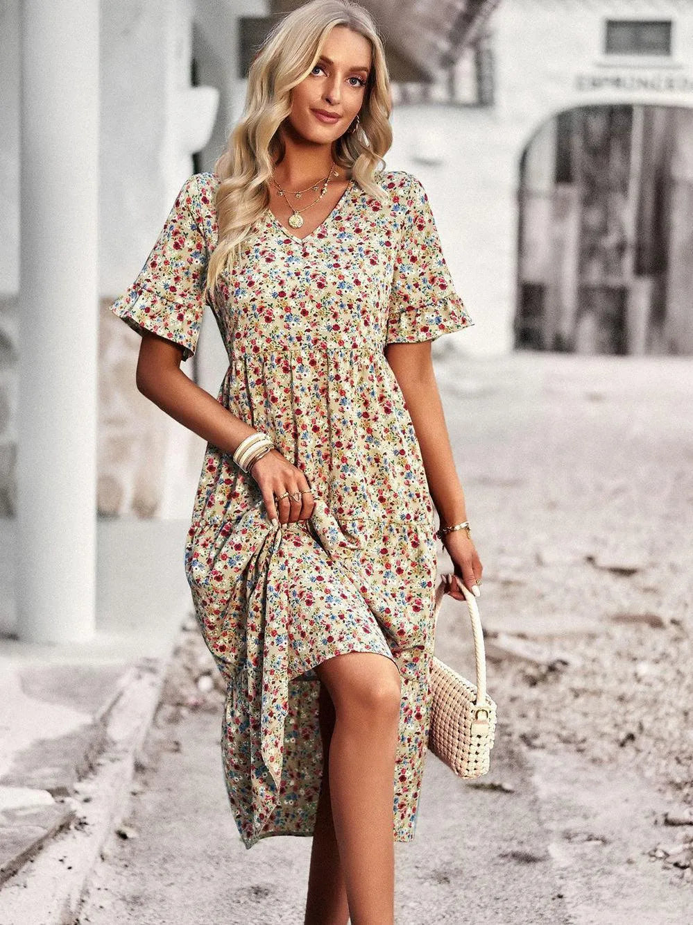 Blonde Frau in geblümtem Sommerkleid, hält eine geflochtene Handtasche, steht auf gepflastertem Weg. Modetrend, Sommermode, Damenkleid.