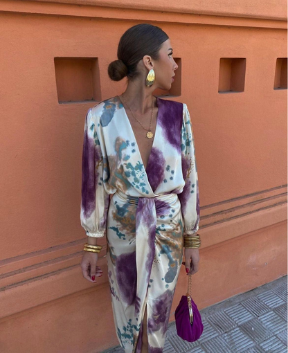 Frau in elegantem, gemustertem Kleid mit goldenen Accessoires und lila Tasche vor orangefarbener Wand. Modetrends, Damenmode, Streetstyle.