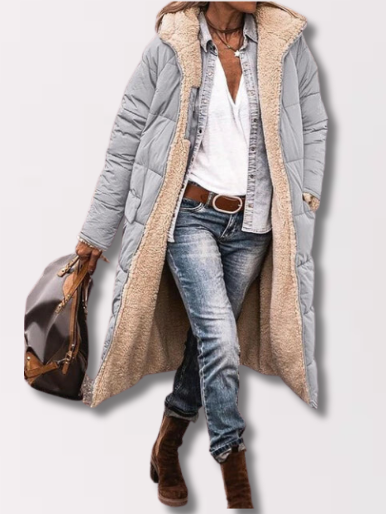 Frau in grauem, langem Steppmantel mit Teddyfutter, Jeans und braunen Stiefeln, hält eine braune Tasche. Wintermode, lässiger Stil.